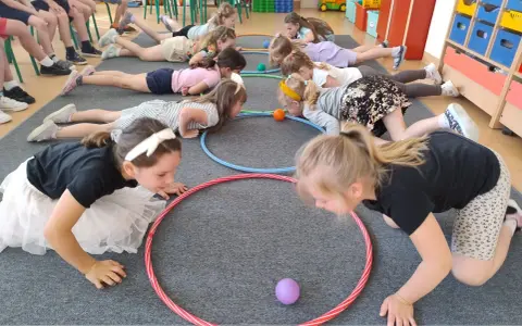Dzieci leżą na podłodze, próbując przesunąć piłeczki za pomocą hula hop. To ciekawe wyzwanie hula hop!