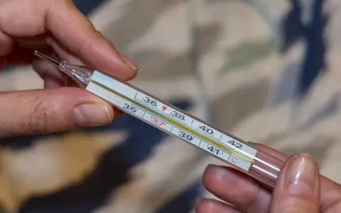 Ręce trzymają termometr, na którym widnieje utrzymująca się temperatura 37 stopni Celsjusza.