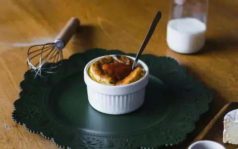 Delicioso souffle de vainilla con salsa de fresa, listo para disfrutar. A su lado, un batidor y un vaso de leche.