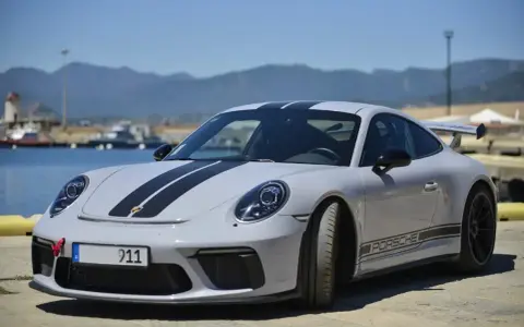 Szary Porsche 911 Carrera z czarnymi pasami na masce i dachu, zaparkowany nad wodą z górami w tle.