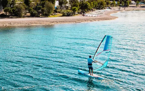 Mężczyzna na desce z żaglem pływa po turkusowej wodzie. Na brzegu widać plażę i drzewa.