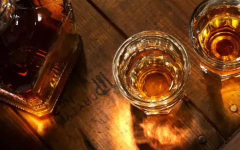 Butelka i dwa kieliszki z bursztynowym płynem, przypominającym koniak lub whisky, stoją na drewnianym blacie.