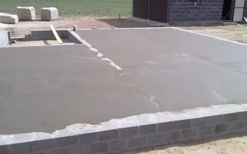 Świeżo wylany beton na chudziaka tworzy fundament pod budowę. W tle widać materiały budowlane i fragment budynku.
