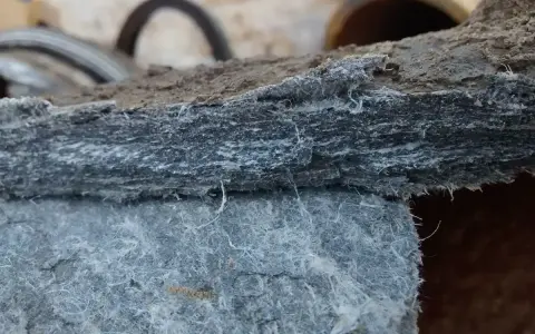 Warstwowa struktura materiału, przypominająca zbite włókna, pokazuje jak wygląda azbest. Widać drobne, białe nitki wystające z szarej masy.