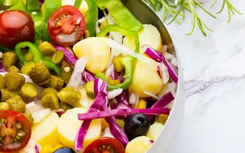 Ensalada campera vibrante con patatas, tomates, pepinillos, pimientos verdes, cebolla morada y aceitunas negras en un bol metálico.