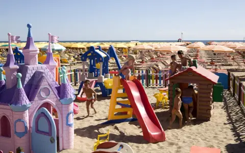 Bambini giocano in un'area giochi colorata sulla sabbia, con castello e scivoli, mentre ombrelloni gialli e lettini creano un'atmosfera da **sunday beach Rimini**.