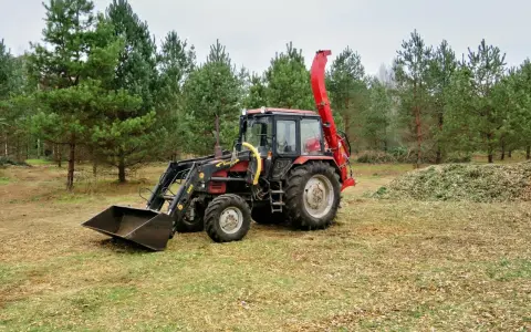 Czerwony ciągnik z ładowaczem i zamontowanym z tyłu rębak do drewna ciągnikowy, gotowy do pracy w lesie.