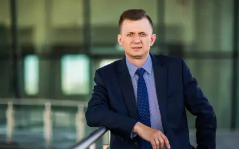Mężczyzna w garniturze i krawacie opiera się o barierkę.