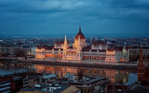 Budynek Parlamentu w Budapeszcie nocą, odbijający się w Dunaju. Miasto tętni życiem, aspirując do miana najbardziej imprezowych miast w Europie.