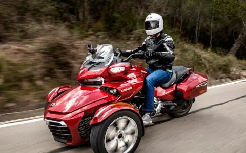 Czerwony pojazd trójkołowy z kierowcą w kasku i kurtce, jadący po krętej drodze. Idealny dla tych, którzy szukają wrażeń, ale nie mają prawa jazdy na motocykl.