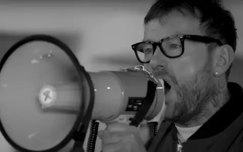 Un homme avec des lunettes et un mégaphone, sa voix résonne. Le groupe est en plein concert.
