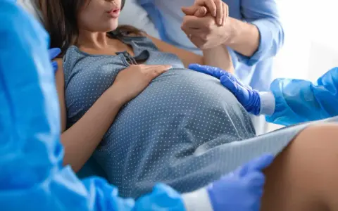 Donna in travaglio, mani che si stringono, personale sanitario con guanti blu. Si percepisce la tensione e l'attesa per lo scollamento membrane.