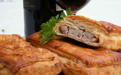 Pâté en croûte, une spécialité lorraine savoureuse, avec une garniture de viande visible. Une bouteille de vin rouge et du persil complètent la scène.