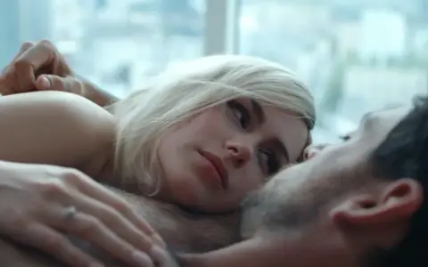 Blondynka i brunet leżą blisko siebie, ich ciała stykają się. Intymna scena, która sugeruje seks na scenie.