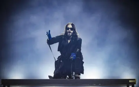 Jared Leto w niebieskich rękawiczkach, w okularach i długim płaszczu, klęczy na scenie. Jego zespół pewnie czeka na jego znak.