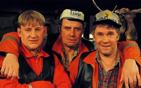 Tre män i orangea jackor och kepsar, en av dem med en uppstoppad älg i bakgrunden. Rollistan i Pistvakt.