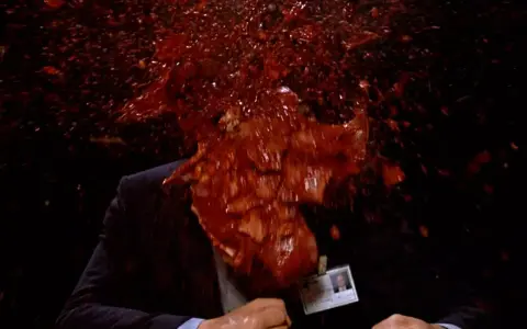 Un uomo in giacca e cravatta, vittima di un evento cruento, con sangue e detriti che esplodono in un'immagine tipica di un genere cinematografico con scene truculente.