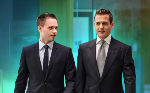 Due uomini in abiti eleganti, stile Harvey Specter, camminano in un ufficio moderno.