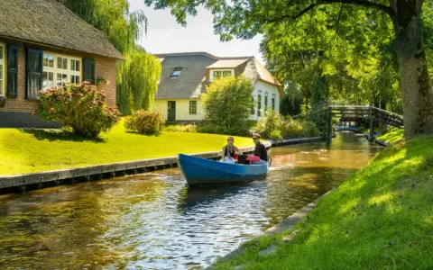 Ein Paar fährt mit einem blauen Boot durch einen Kanal im "Klein Venedig Holland". Reetgedeckte Häuser und grüne Ufer säumen den Weg.