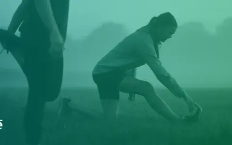 Två personer stretchar utomhus i dimma. En kvinna på knä sträcker sig mot sin fot. Detta kan vara en del av rehab benhinneinflammation.