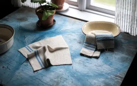 Två virka handdukar med blå ränder ligger på ett blått bord. En krukväxt och en skål syns också.