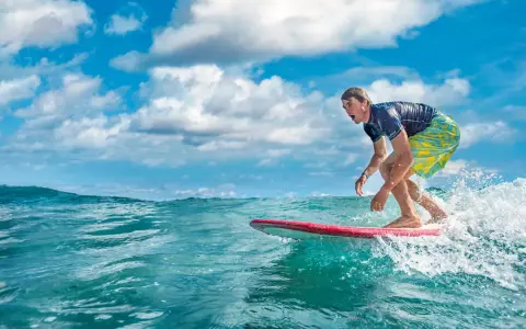 Ein Surfer reitet eine Welle vor der Küste der Dominikanischen Republik. Der Mann trägt ein blaues Shirt und bunte Badeshorts.