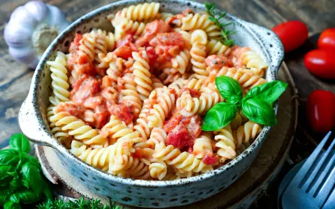 Fusilli-Nudeln in cremiger Tomaten-Sahne-Sauce, garniert mit frischem Basilikum. Daneben liegen Kirschtomaten und Knoblauch.