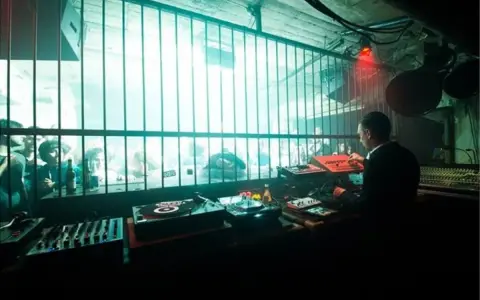 DJ set in un locale underground, luci al neon e folla che balla dietro le sbarre. Atmosfera da berlino rave.