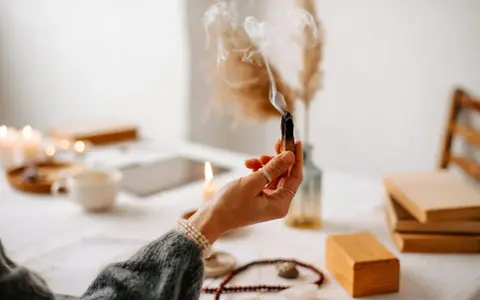 Mano sosteniendo un palo santo humeante, rodeada de velas, libros y cristales, creando un ambiente de sanación y paz.