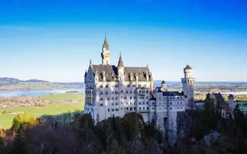 Sagolikt slott i Tyskland, Neuschwanstein, reser sig majestätiskt på en klippa med en sjö och grönskande landskap i bakgrunden.