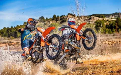 Dos pilotos de enduro compiten en un terreno embarrado, uno con motor 2t y otro 4t, salpicando agua.