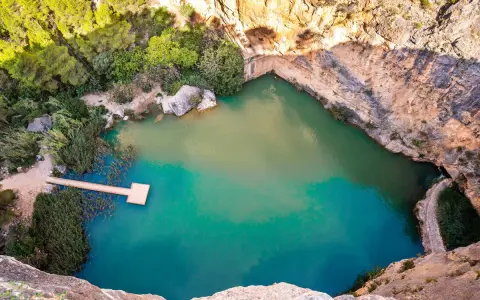 Un **charco azul** en Chiva, rodeado de rocas y vegetación. Un muelle de madera se adentra en sus aguas turquesas.