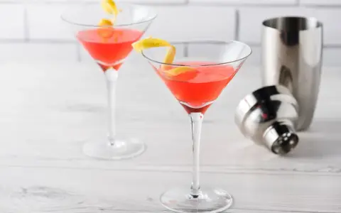 Dwa drinki Cosmopolitan z plasterkiem cytryny, obok metalowa shakera. Idealny przepis na Cosmopolitan drink.