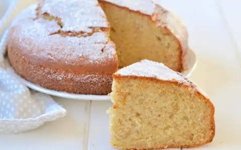 Una fetta di soffice torta di banane mature, spolverata di zucchero a velo, accanto alla torta intera su un piatto bianco.