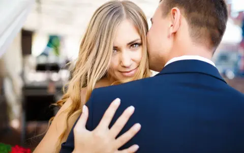 Blondynka z zielonymi oczami patrzy w obiektyw, gdy mężczyzna w granatowym garniturze, numerologiczna 8 mężczyzna, całuje ją w policzek.