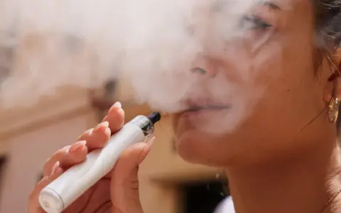 Una mujer exhala vapor de un vaper blanco. El vapor parece rodear su rostro, sugiriendo que el vaper encharca los pulmones.