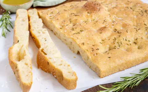 Frisch gebackene Focaccia, ein Klassiker unter den italienischen Brotsorten, mit grobem Salz und Rosmarin bestreut. Daneben liegen zwei geschnittene Stücke.
