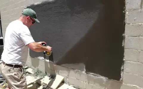 Un homme utilise une ponceuse pour araser un mur extérieur fraîchement enduit.