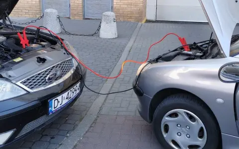 Dwa samochody z otwartymi maskami, połączone kablami rozruchowymi. Pomoc w awaryjnej sytuacji, jak sprawdzić masę w aucie.