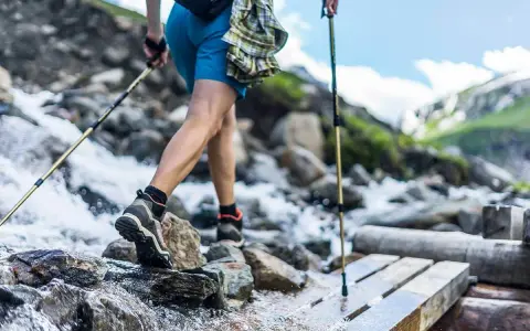 Une personne en tenue de ppg sport traverse un ruisseau sur un pont de bois, bâtons de randonnée en main. Paysage de montagne.
