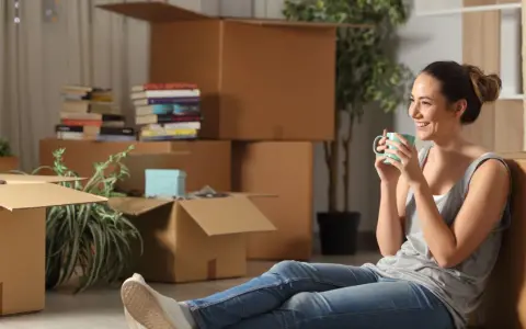 Une femme boit une tasse de café, entourée de nombreux cartons de déménagement.