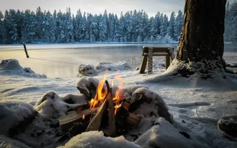 Ein Lagerfeuer wärmt beim Zelten im Winter am gefrorenen See. Schnee bedeckt die Ufer und den Wald im Hintergrund.
