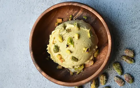 Cremoso helado de pistacho servido en un cuenco de madera, adornado con trozos de pistacho y acompañado de más pistachos enteros esparcidos alrededor.