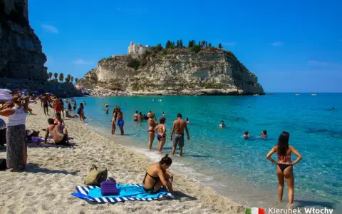 Słoneczna plaża z turkusową wodą i klifem, na którym stoi kościół. Ludzie kąpią się i opalają, ciesząc się jednymi z najpiękniejszych plaż we Włoszech.