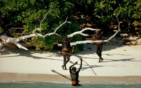 Trzech mężczyzn z plemienia Sentinelese, uzbrojonych w łuki i strzały, stoi na plaży. Sentinel Island, ich dom, chroni ich przed światem.