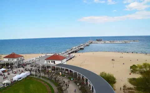 Sopot beach z molo, kawiarniami i spacerującymi ludźmi. W oddali widać statki.