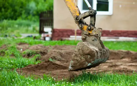 Błotnista łyżka koparki wykonuje niwelację terenu wokół domu.