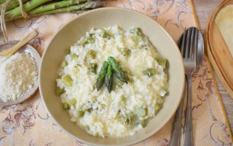 Kremowe risotto ze szparagami, posypane parmezanem, z trzema zielonymi szparagami na wierzchu. Obok miseczka z tartym serem i pęczek świeżych szparagów.