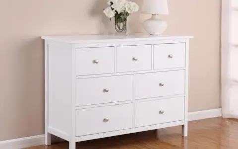 Commode blanche 3 tiroirs, prête à être relookée. Un vase de fleurs et une lampe complètent la scène.