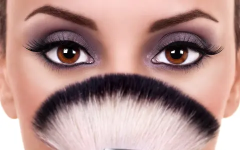 Occhi grandi e tondi esaltati da un trucco smokey viola e ciglia finte, con un pennello da trucco in primo piano.